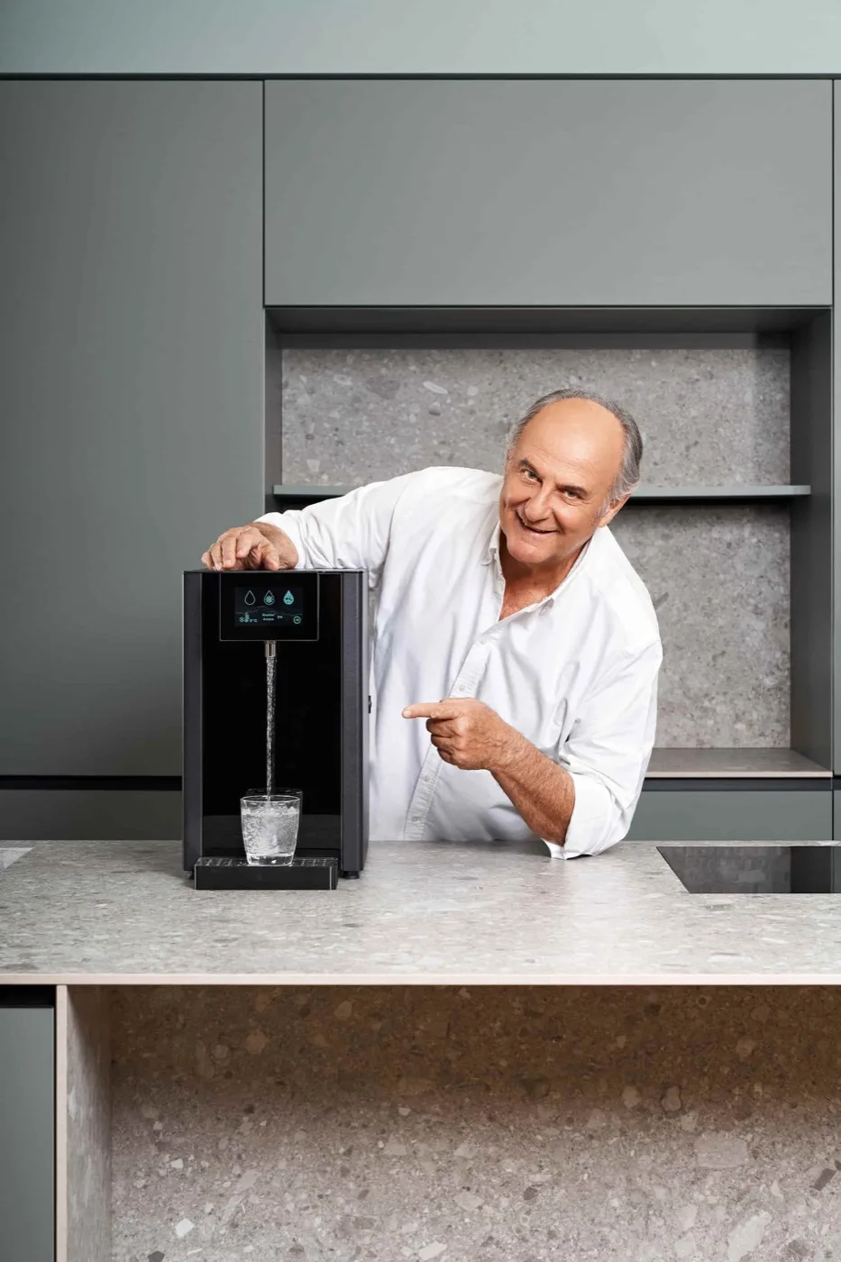 Gerry Scotti accanto a un depuratore Aquamed che riempie un bicchiere, in cucina